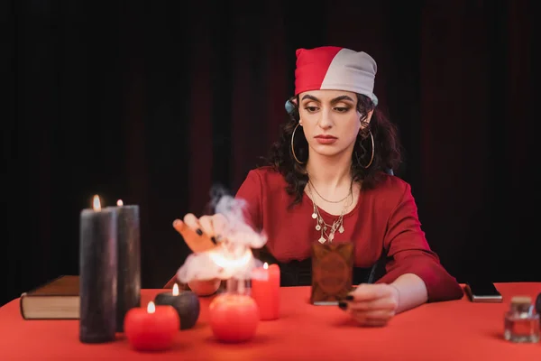 Gypsy medium holding tarot card near blurred candles on table on black — Stock Photo