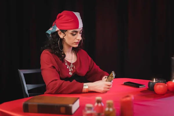 Gypsy fortune teller holding tarot cards near blurred book and witchcraft supplies isolated on black - foto de stock