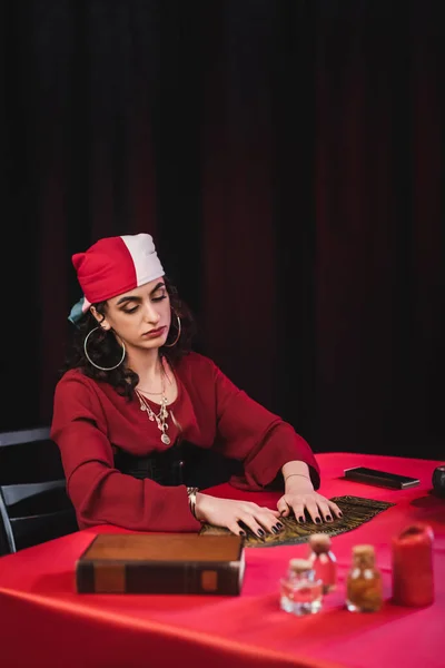 Gypsy fortune teller touching tarot cards near book and jars on table isolated on black — Foto stock