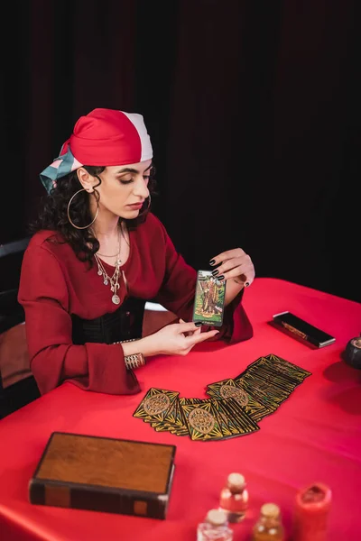KYIV, UKRAINE - FEBRUARY 23, 2022: Young gypsy fortune teller holding tarot card near jars and book isolated on black — Stock Photo