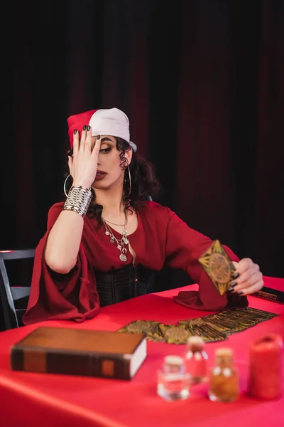 Scared gypsy medium holding tarot card near book on black — Photo de stock