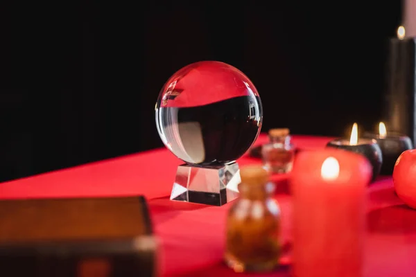 Glass orb near blurred candles and witchcraft supplies on table isolated on black — Stockfoto