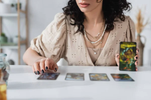 Vista recortada del adivino sosteniendo cartas borrosas del tarot cerca de la mesa - foto de stock