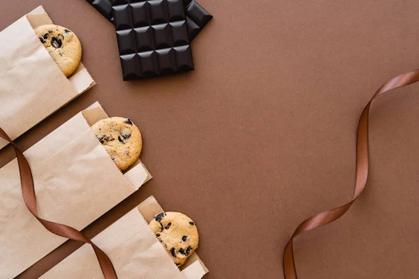 Draufsicht auf Kekse in Bastelverpackungen, Schokoriegel und Schleife auf braunem Hintergrund — Stockfoto