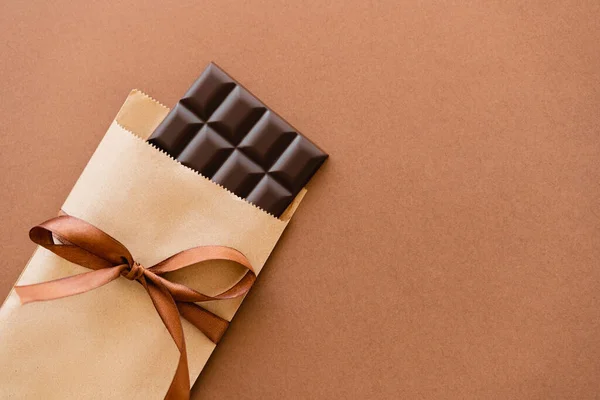 Top view of dark chocolate bar in craft package with ribbon on brown background — Stockfoto