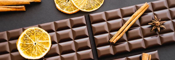 Top view of cinnamon and dry orange sticks on chocolate bars on black background, banner — Stockfoto