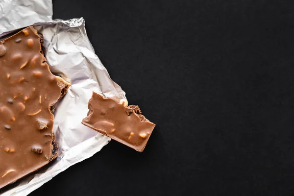 Top view of milk chocolate with nuts on foil on black background — Stock Photo