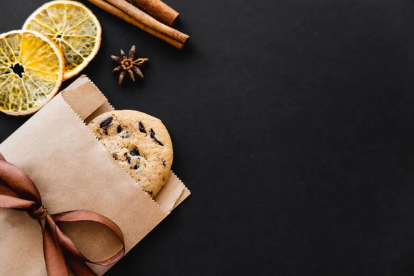 Top view of cookie in craft package, dry orange slices and spices on black background — Photo de stock
