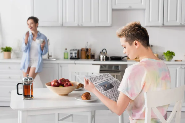 Young pangender person reading newspaper near partner with tea cup - foto de stock