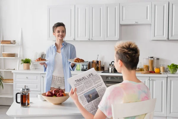 Alegre pansexual pessoa segurando croissants perto do parceiro com viagem vida jornal — Fotografia de Stock