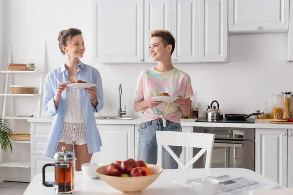 Happy pangender couple with tasty croissants looking at each other near blurred teapot and apples — Stock Photo