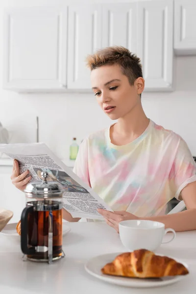 Bigender person with short hair reading morning newspaper near teapot and blurred croissants - foto de stock