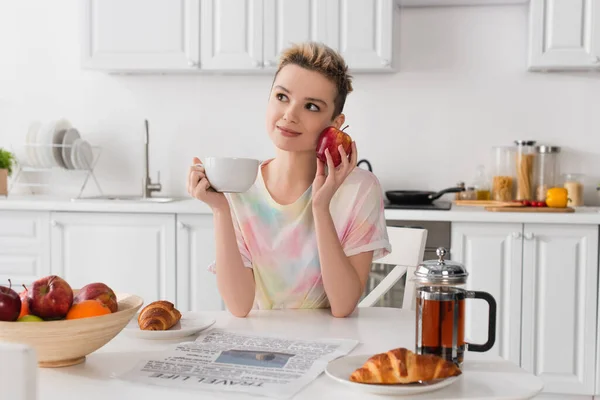 Persona pansessuale sorridente con mela e tazza di tè seduta vicino a cornetti e giornali — Foto stock
