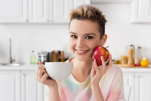 Щаслива психічна людина з чашкою чаю і свіжим яблуком, дивлячись на камеру на кухні — стокове фото