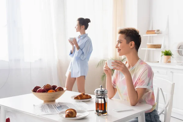 Smiling pansexual person sitting with cup of tea near croissants and partner on blurred background — Stock Photo