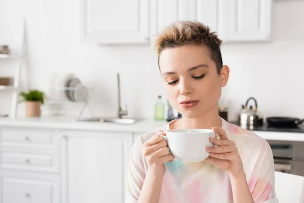 Young bigender person with short hair holding cup of tea in blurred kitchen — Photo de stock