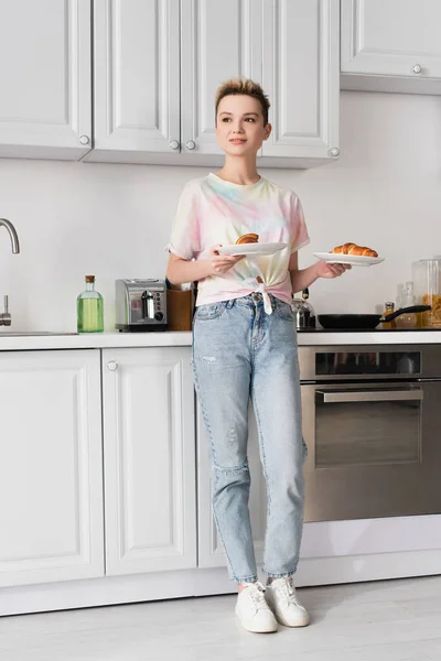 Full length of positive pangender person in jeans holding tasty croissants in kitchen — стоковое фото
