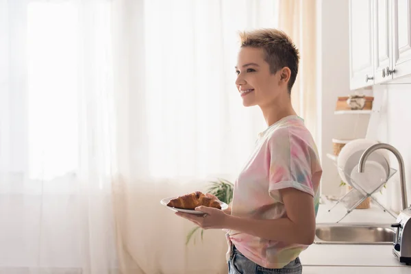 Вид сбоку счастливого бигендера с короткими волосами, держащими тарелку с круассаном на кухне — стоковое фото