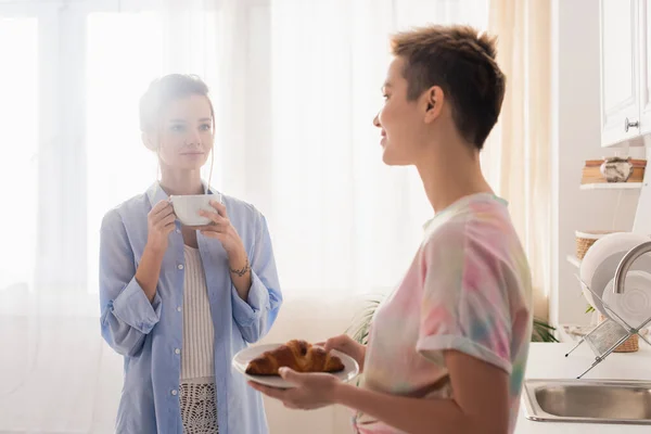 Bigender couple with croissant and tea cup looking at each other in kitchen - foto de stock