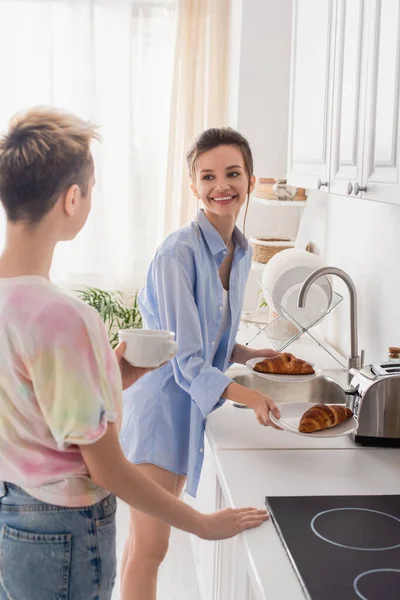 Feliz pansexual persona sosteniendo croissants cerca pareja con taza de té - foto de stock