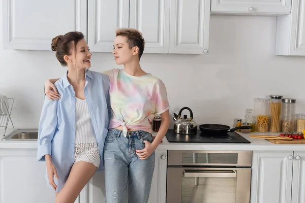 Happy bigender couple looking at each other near stove in kitchen — стоковое фото