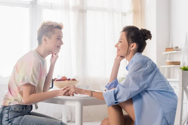 Side view of bigender couple holding hands and smiling at each other while sitting in kitchen - foto de stock