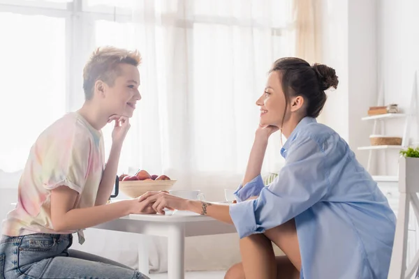 Side view of bigender couple holding hands and smiling at each other while sitting in kitchen — Photo de stock