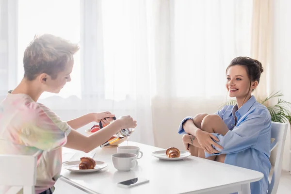 Pangender person pouring tea near happy partner and delicious croissants - foto de stock