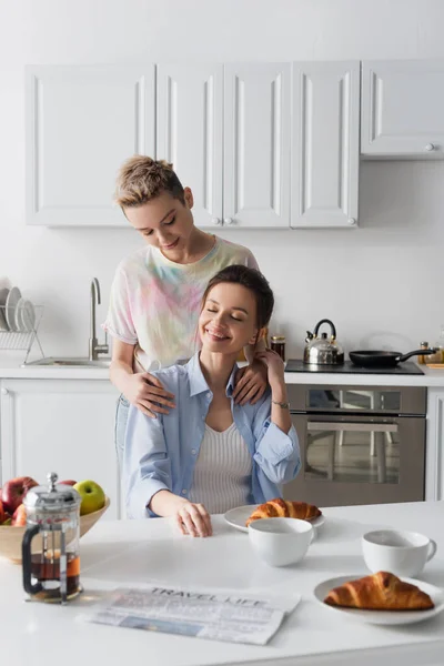 Pansexual person hugging shoulders of smiling partner sitting with closed eyes near breakfast — Stock Photo
