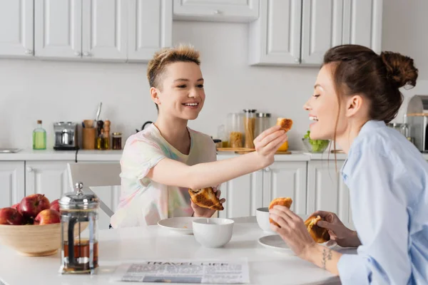 Smiling bigender person feeding partner with croissant during breakfast in kitchen - foto de stock
