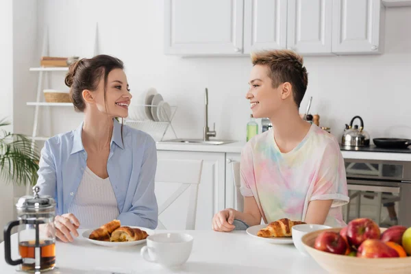 Happy pangender people looking at each other near croissants, tea and blurred apples - foto de stock