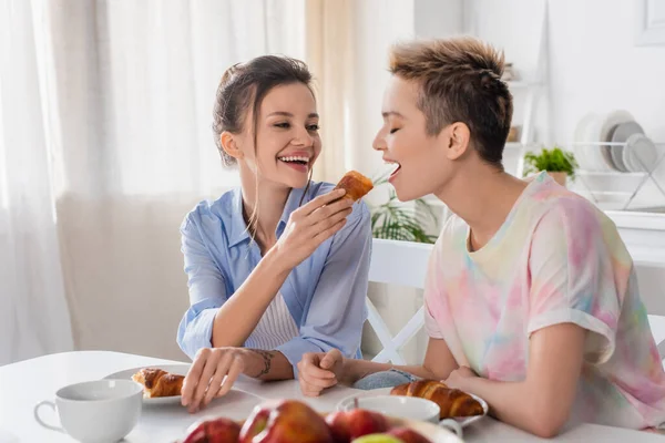 Cheerful pangender person feeding lover with croissant while having breakfast in kitchen — Stock Photo