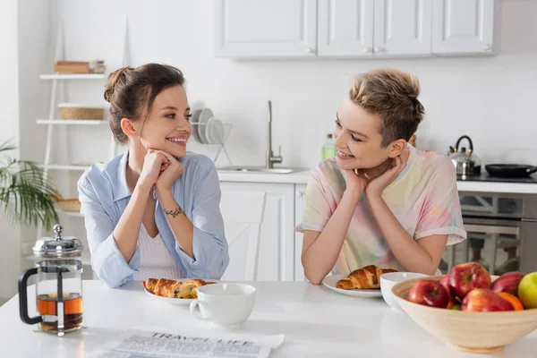 Pleased pangender couple looking at each other near croissants, tea and blurred apples — Fotografia de Stock