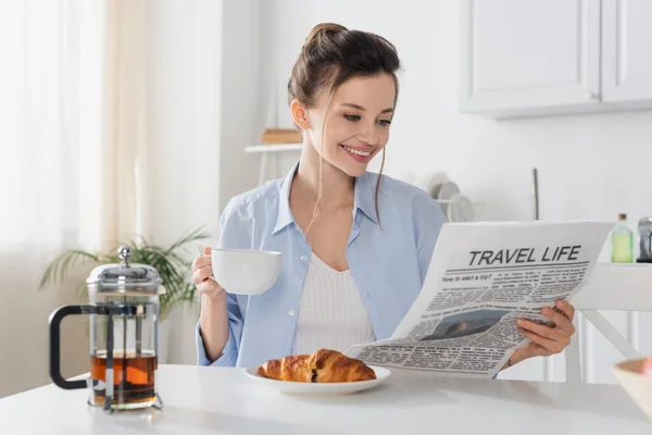 Mulher sorrindo com xícara de chá lendo jornal de viagem vida perto de croissant delicioso — Fotografia de Stock