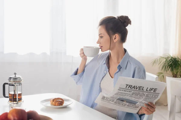 Young woman with travel life newspaper drinking tea near tasty croissant — Photo de stock
