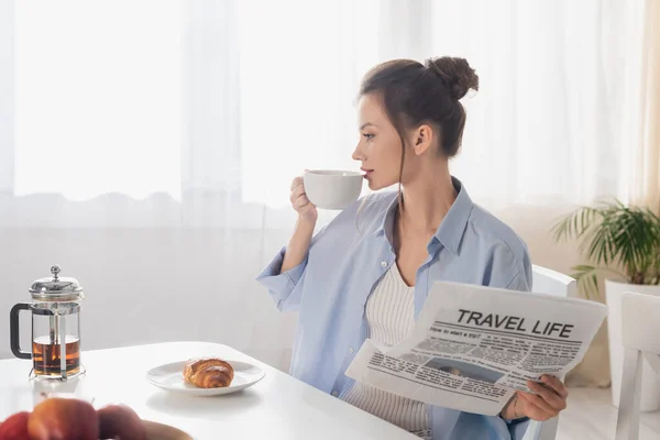 Mujer joven con la vida de viaje periódico beber té cerca de croissant sabroso - foto de stock