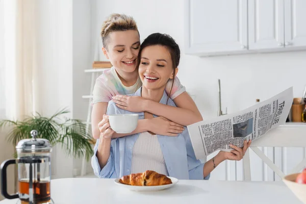 Glückliche pansexuelle Person umarmt lächelnden Partner sitzt mit Zeitung und Teetasse — Stockfoto