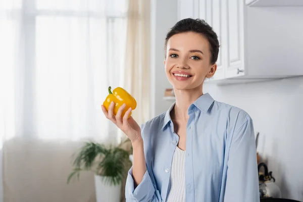 Happy brunette woman with fresh bell pepper smiling at camera in kitchen — Stockfoto