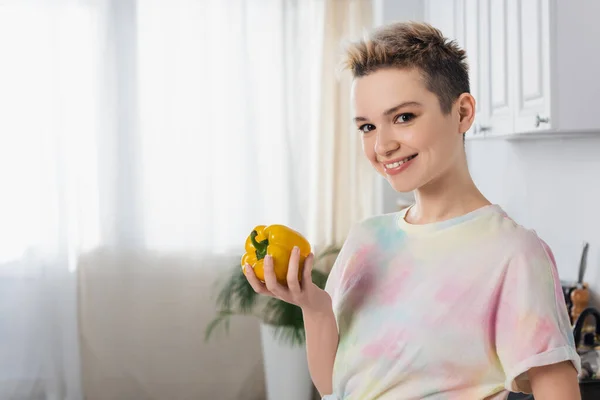 Happy pangender person holding whole bell pepper and looking at camera in kitchen - foto de stock