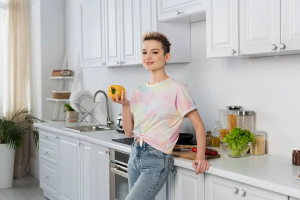 Bigender person with short hair smiling at camera while holding whole bell pepper in kitchen — Photo de stock