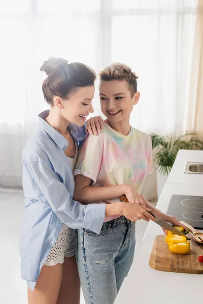 Cheerful pangender person helping partner cutting bell pepper in kitchen — Stock Photo