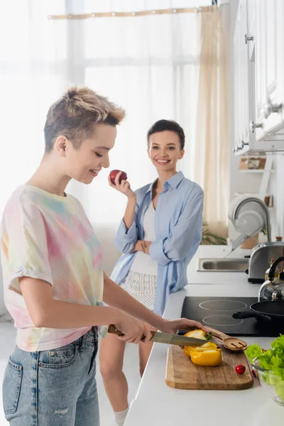 Cheerful bigender person holding apple near lover cutting vegetables in kitchen — стоковое фото