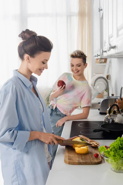 Blurred pansexual person cutting bell pepper near partner with fresh apple in kitchen — Stock Photo