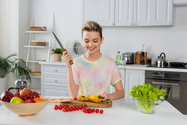 Glückliche zweigeschlechtliche Person mit Messer, die neben Paprika, Kirschtomaten, Salat und Früchten in der Küche sitzt — Stockfoto