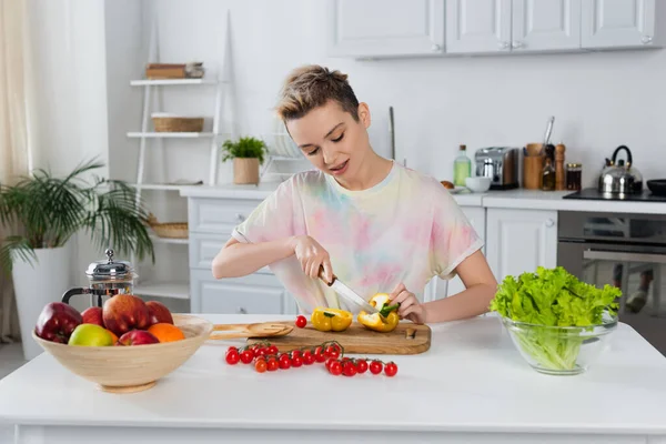 Jovem bigender pessoa corte pimentão perto de alface, tomate cereja e frutas — Fotografia de Stock