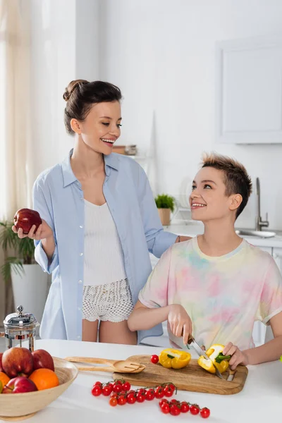 Happy bigender person holding apple near partner cutting vegetables in kitchen — Stock Photo