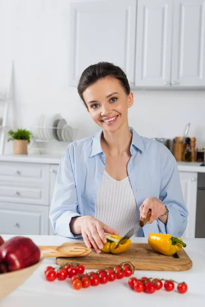 Joyful woman cutting bell pepper near fresh cherry tomatoes in kitchen — Stock Photo