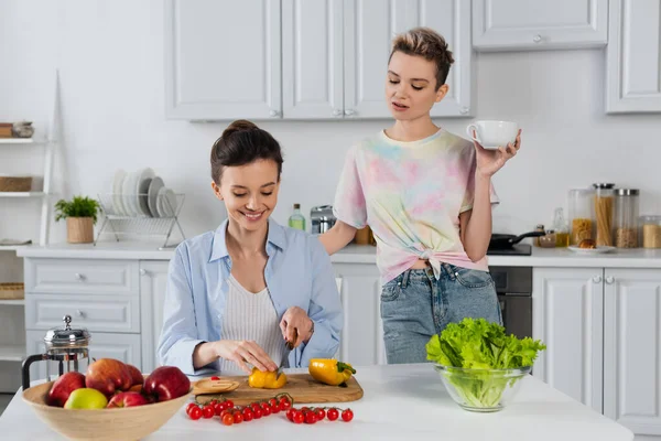 Smiling pansexual person cutting bell pepper near fresh vegetables and partner with tea cup - foto de stock