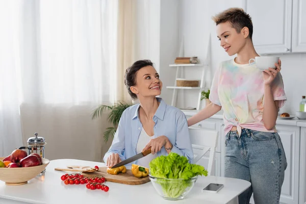 Cheerful pangender person cutting fresh vegetables near lover with cup of tea — стоковое фото