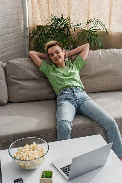 Glückliche bigeschlechtliche Person entspannt sich auf dem Sofa und schaut Film auf Laptop in der Nähe Schüssel Popcorn — Stockfoto